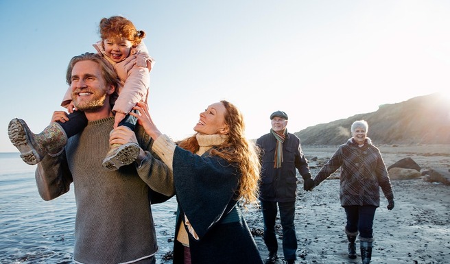 A happy family walks along a beach at sunset. A man carries a smiling child on his shoulders, while a woman stands beside them, smiling. In the background, an older couple holds hands, enjoying the scene. The atmosphere is joyful and warm.
