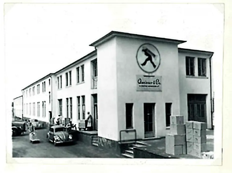 En sort-hvid foto af en bygning med en stor logo af en person i bevægelse på facaden. Der står kasser foran bygningen, og der er nogle personer og biler i nærheden. Bygningens stil ser ud til at være fra midten af det 20. århundrede.