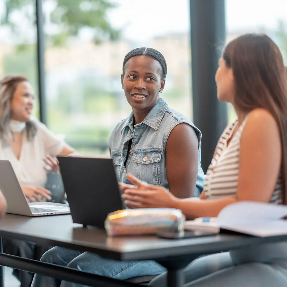 En gruppe mennesker sidder ved et bord i et lyst rum, hvor de diskuterer og arbejder på laptops. Den centrale person smiler og ser engageret ud. Det er en afslappet atmosfære med fokus på samarbejde.