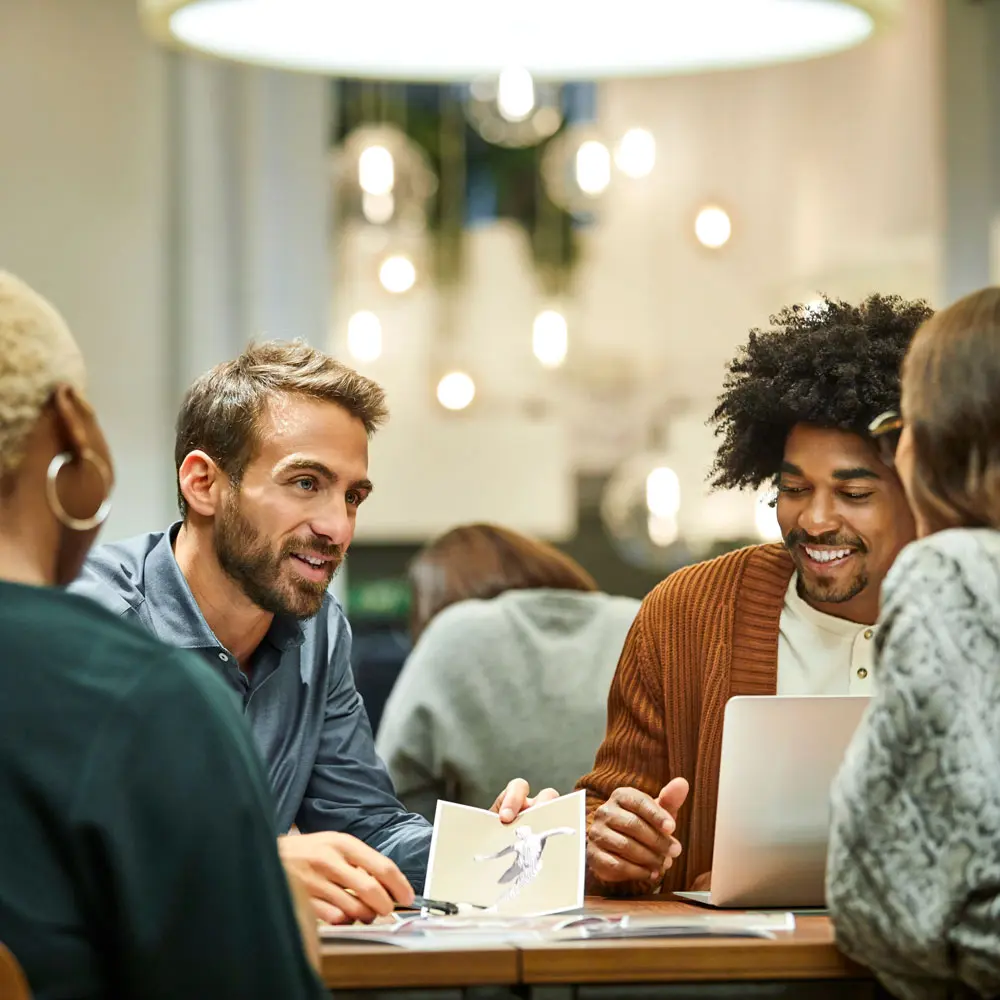 Tre personer sidder omkring et bord og diskuterer. En mand med kort hår og skæg smiler, mens han peger på et billede. En anden mand med krøllet hår lytter opmærksomt. Atmosfæren er uformel og inspirerende.