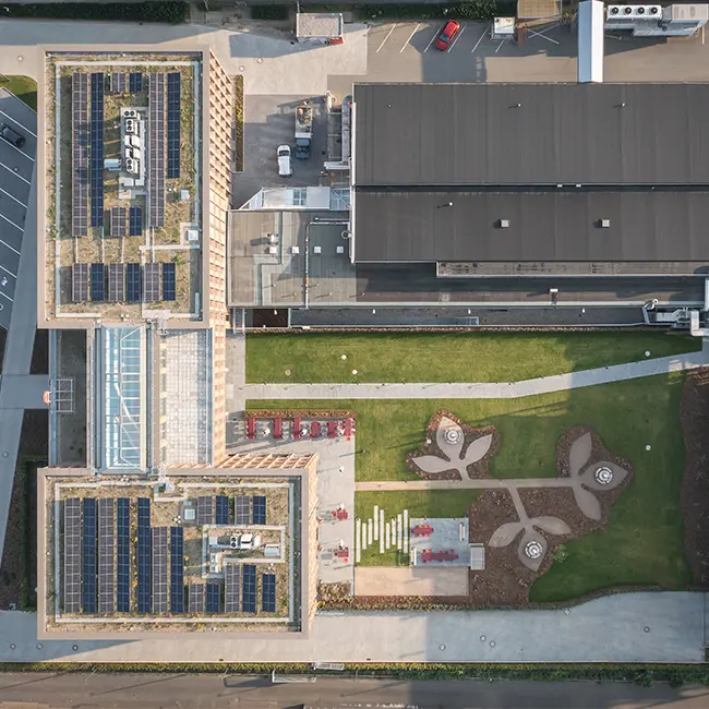 Luftfoto af to bygninger, der har solpaneler på taget. Under bygningerne er der en grøn græsplæne med stier og et kunstnerisk landskab designet med stensætninger og små siddepladser. Parkeringspladser ses til venstre.