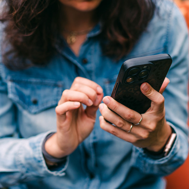En person i en blå skjorte bruger en smartphone, mens de fokuserer på skærmen med en hånd. Den anden hånd berører skærmen. Baggrunden er uskarp, hvilket fremhæver personen og telefonen.