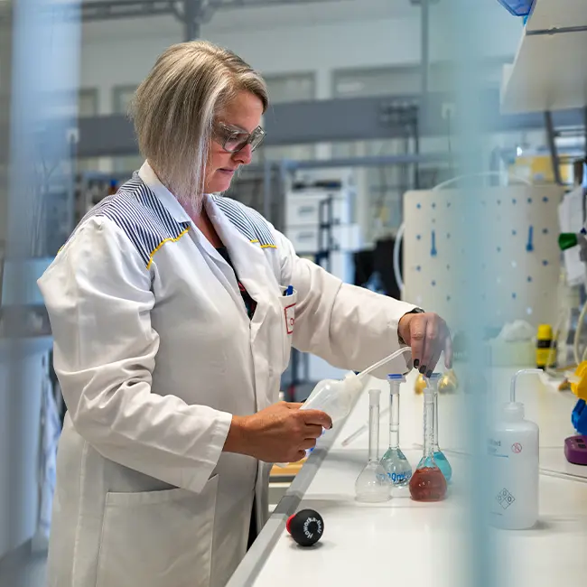 En kvinde i laboratoriekittel arbejder med reagenser i et kemilaboratorium. Hun bruger en pipette til at overføre væske til et glasudstyr. Baggrunden viser laboratorieudstyr og flasker.