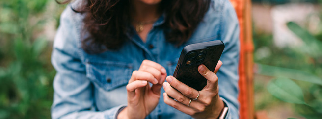 En person sidder med en smartphone i hånden og bruger den, mens de er omkranset af grønne planter. Personen har krøllet hår og bærer en denimjakke.