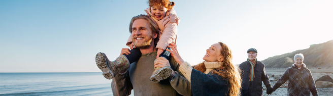 A happy family enjoys a sunny day at the beach. A man carries a young girl on his shoulders, while a woman smiles beside them. In the background, an older couple strolls along the shore. The scene conveys joy and togetherness.